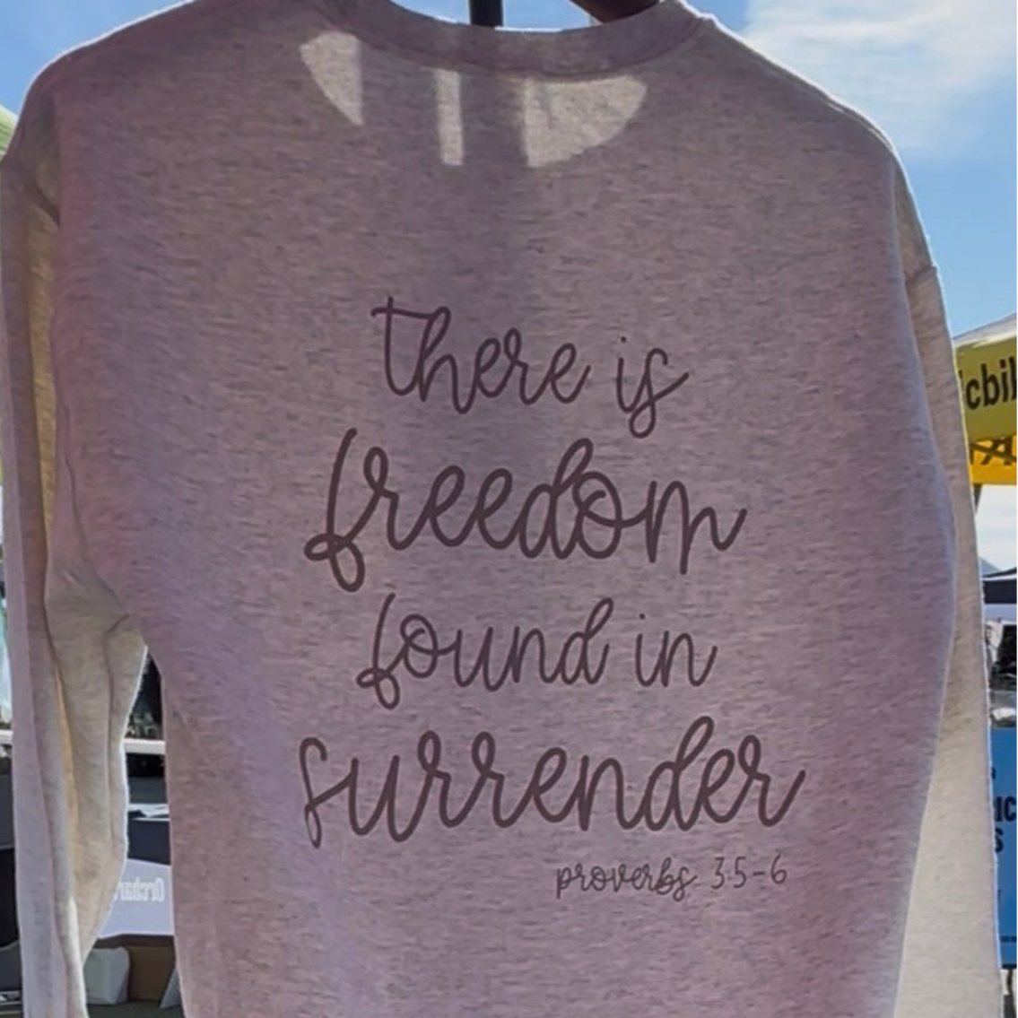 Light gray sweatshirt with 'there is freedom found in surrender' text on a blurred outdoor background