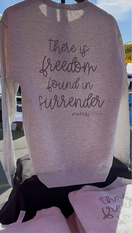 Light gray sweatshirt with 'there is freedom found in surrender' text on a blurred outdoor background